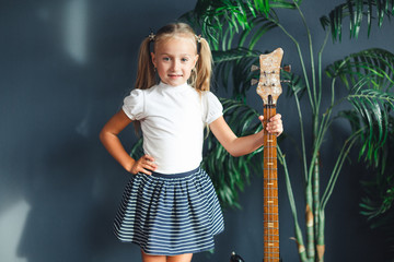 young blonde girl with tails in white t-shirt, skirt and sandals with electric guitar at home looking at camera and smiles.