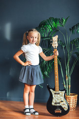 young blonde girl with tails in white t-shirt, skirt and sandals with electric guitar at home looking at camera and smiles.