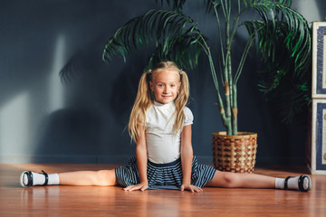 Beautiful little blonde girl with hair gathered in tails, white t-shirt, white socks and gray skirt sitting on twine at home looking at camera and smiles.