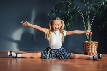 A little girl with hair gathered in tails, white t-shirt, white socks and gray skirt, adorable young talented dancer does ballet poses and stretching exercises on the floor at home