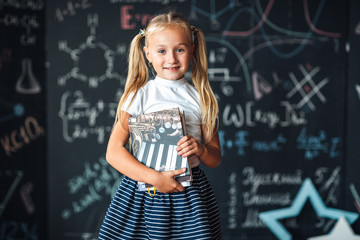 Smiling little blonde girl with hair gathered in tails, white t-shirt, and gray skirt is holding book in a classroom with a сhalkboard covered with formulas and figures.