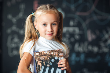 Smiling little blonde girl with hair gathered in tails, white t-shirt, and gray skirt is holding book in a classroom with a chalkboard covered with formulas and figures.