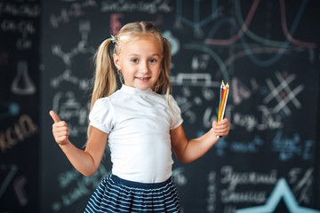 Cute girl in school uniform with colored pencils. Go to school for the first time. Girl indoors of the class room with blackboard on a background shows the thumb. Back to school