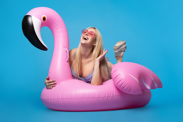 Picture of blonde in bathing suit and in pink glasses lying on pink inflatable flamingo on empty blue background © Sergey Mironov