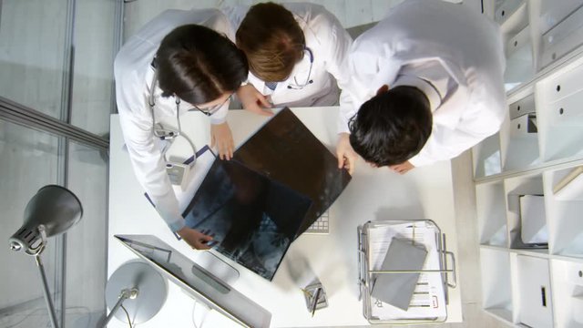 Top view shot of physicians in white coats walking up to male colleague working on computer in office. They are showing MRI and x-ray of patient and asking for advice