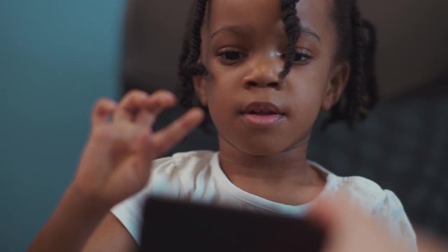 Pretty excited little afro-american kid girl using a smartphone taking pictures indoors. Close-up of beautiful cute african child smiling and playing with a phone.