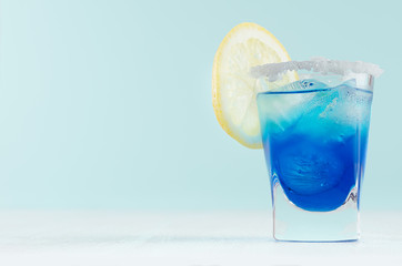 Blue curacao cocktail with lemon slice, ice cubes, sugar rim in shot glass on elegant white wood...
