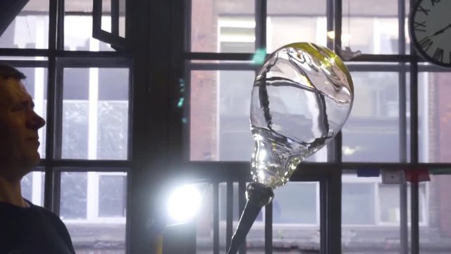 Glassblower looking at a glass product in front of a window. Close-up.