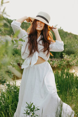 Beautiful carefree long hair asian girl in white clothesand straw hat  on river side in the grass at sunset. Sensitivity to nature concept