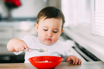 Funny little girl in the kitchen during the day eating oatmeal porridge from a red plate all dirty