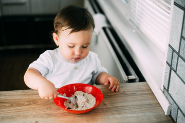 Funny little girl in the kitchen during the day eating oatmeal porridge from a red plate all dirty