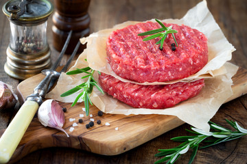 Raw mince meat beef burgers on a wooden cutting board with fresh rosemary, cloves of garlic, pepper corns and salt