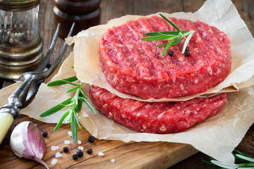 Raw mince meat beef burgers on a wooden cutting board with fresh rosemary, cloves of garlic, pepper...