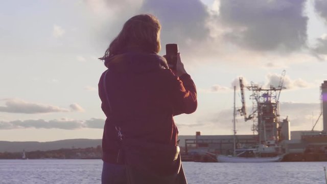4K anamorphic video shot with gimbal of a young Norwegian woman taking photo of a beautiful sunset with her smartphone at the Soerenga pier, a popular tourist destination in Oslo, Norway.