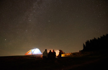 Night camping in the mountains near forest. Group of four happy friends hikers sitting together around bonfire beside glowing tourist tent. On background night starry sky full of stars. Back view