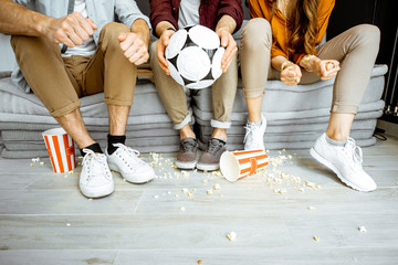 Friends watching football match. View on their legs with pop cornes and ball on the floor