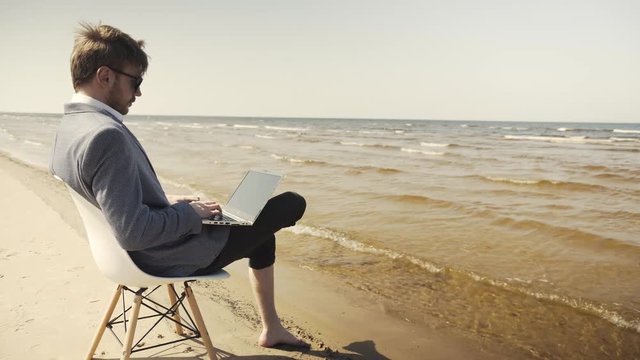 young business man in suit sitting in a beach area, working with laptop pc on his lap. Sea and beach in background.