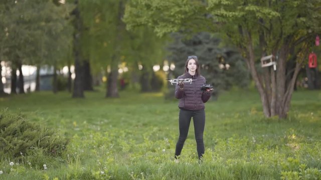 Drone With Camera Landing in Hand of Young Caucasian Female With Remote Controller in Green Field of Public Park. Blurred Background