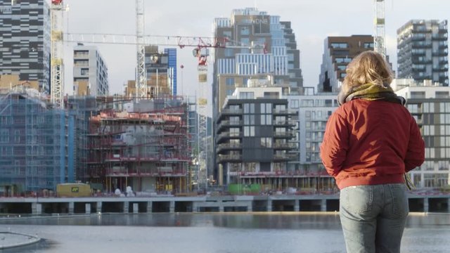 4K anamorphic video with forward motion of a young Norwegian woman with washed out jeans and a red jacket looking at the construction of buildings and architecture in Bjoervika in Oslo, Norway.