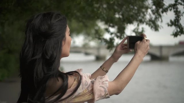 Close Up of an attractive Hispanic tourist taking a picture of the Thames in London from a river side walk, holding her phone to frame the shot