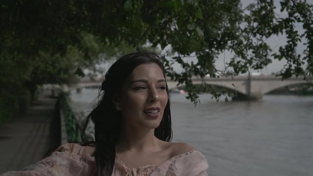 Attractive and smiling Latina tourist taking a selfie while standing on the railing of river Thames overlooking Putney bridge in London, slow motion shot
