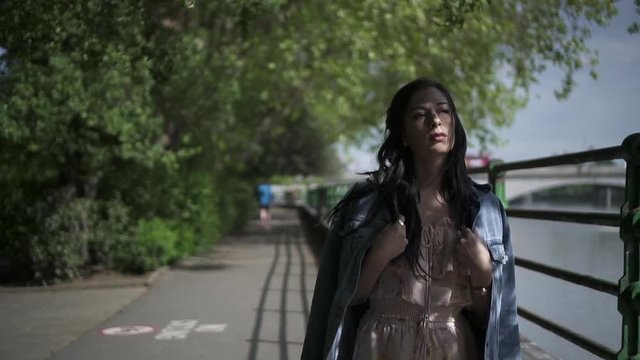 Slow Motion of an attractive latina tourist with black wavy hair and a jean jacket walking in a park in London with a view of Putney bridge behind her