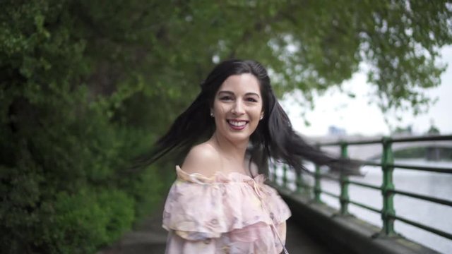 Portrait of an attractive latina tourist with black wavy hair and a floral dress walking in a park in London with a view of Putney bridge behind her