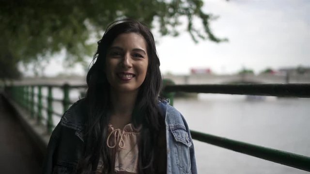 Portrait of an attractive latina tourist with black wavy hair and a jean jacket posing in a park in London with a view of Putney bridge behind her