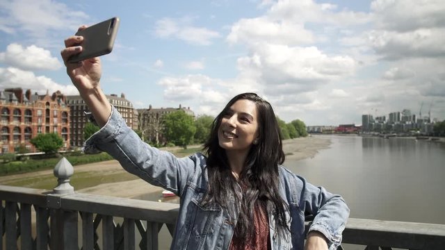 Brunette Latina tourist taking a selfie, posing doing victory sign with her hand, while standing on the railing of a bridge in London