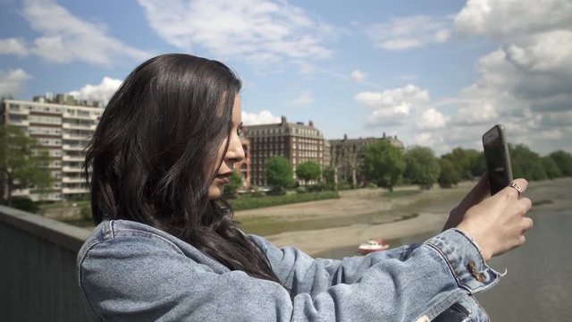 Attractive Hispanic tourist taking a picture of London from a bridge, taking her phone out and holding it to frame the shot