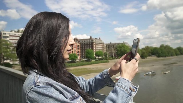 Attractive Hispanic tourist taking a picture of London from a bridge, taking her phone out and holding it to frame the shot