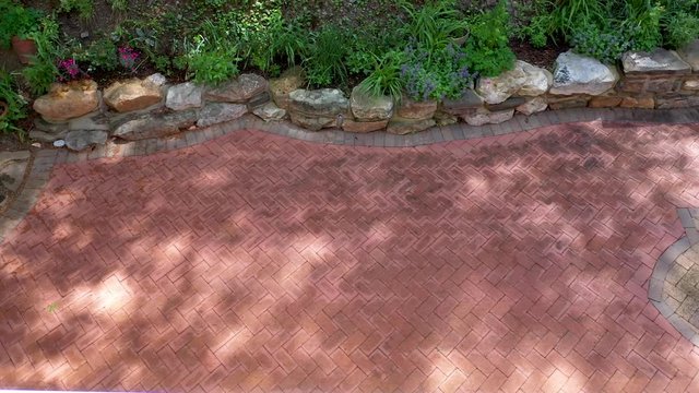 Aerial view looking towards rock wall with gardens merging into forest with herringbone brick pattern on backyard patio.