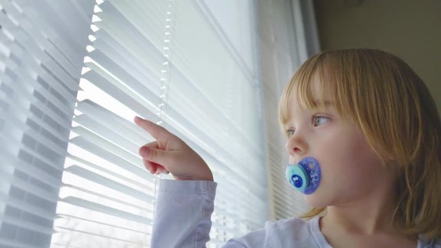 A little girl with a pacifier in her mouth looks out a window and plays with the blinds