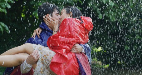 Portrait of young carefree couple in love are wearing protection capes hugging and kissing under the rain on a background of green trees.