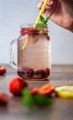 A woman holds in her hand a yellow tube for a cocktail in a glass beaker with a fresh drink of berries and mint on a white background. In the foreground are strawberries,  mint and lemon