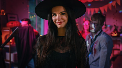 Halloween Costume Party: Gorgeous Young Witch Wearing Dress and Hat Poses seductively. Background: Beautiful She Devil, Scary Death, Count Dracula, Zombie Dancing in the Decorated Room