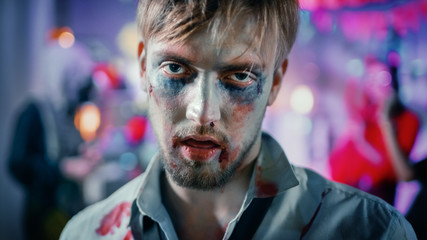 Halloween Costume Party: Portrait of Brain Dead Zombie Wearing Bloody Suit Smiles Creepily. In the Background Monsters Having Fun and Dancing in the Decorated Room