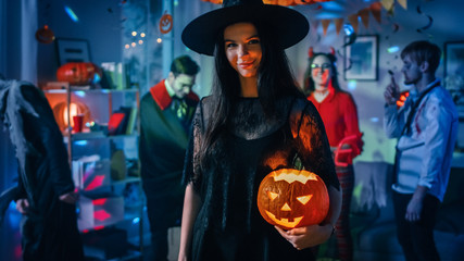 Halloween Costume Party: Gorgeous Seductive Witch Wearing Dress Holds Burning Pumpkin. Background: Beautiful Devil, Scary Death, Count Dracula, Zombie Dancing in the Decorated Room - obrazy, fototapety, plakaty