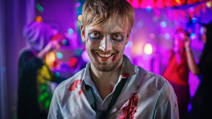 Halloween Costume Party: Portrait of Brain Dead Zombie Wearing Bloody Suit Smiles Creepily. In the Background Monsters Having Fun and Dancing in the Decorated Room