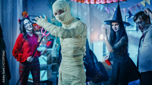 Halloween Costume Party: Old Skinny and Bandaged Mummy Dances. In the Background Zombie, Death, Witch and She Devil Have Fun in a Monster Party Decorated Room