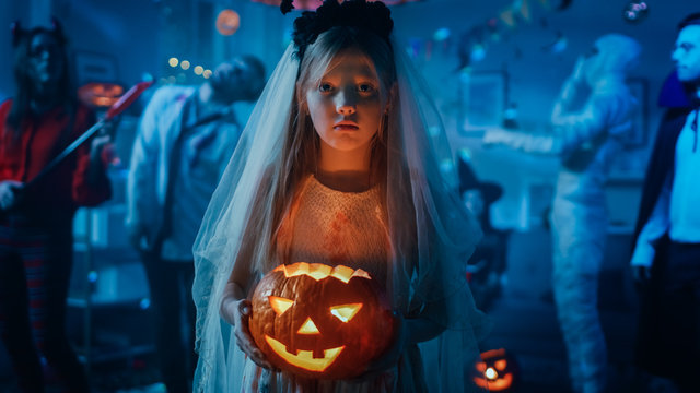 Halloween Costume Party: Little Girl in a Bloody White Bride Dress Holding Scary Doll. Zombie, Blood Thirsty Dracula, Mummy, Bewitching Witch and Dazzling She Devil Having Fun in Decorated Room