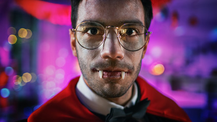 Halloween Costume Party: Close-up Portrait of Count Dracula Smiling Creepily, Shows His Deadly Bloody Fangs. In the Background Thematically Decorated Room with Scary Stuff. Deadly Blue Colors