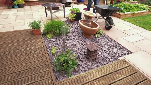 Footage of man working in garden, getting slate out of a wheelbarrow and throwing it on the ground in a new area.  Garden refresh/remake