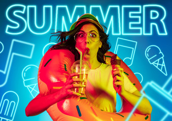 Female model in trendy neon light on blue background. Modern design. Beautiful young woman posing in cap and sunglasses with the drink. Concept of facial expression, summer, vacation, music, open-air.
