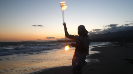 A male fire dancer or performer in silhouette spinning a flaming staff on the beach at sunset with ocean waves in slow motion. - Powered by Adobe