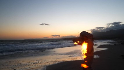 A man in silhouette dancing and spinning a burning fire staff on the beach at sunset with flames and ocean waves SLOW MOTION - Powered by Adobe