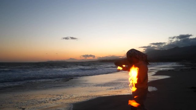 A man in silhouette dancing and spinning a burning fire staff on the beach at sunset with flames and ocean waves SLOW MOTION