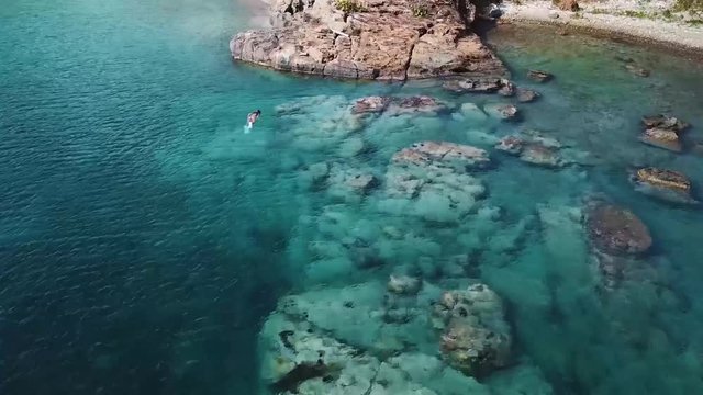 Island ocean swimming and Snorkeling beautiful Millennial girl on vacation swims over fish in biniki filming fish and reef with action camera as drone flies over looking down at her