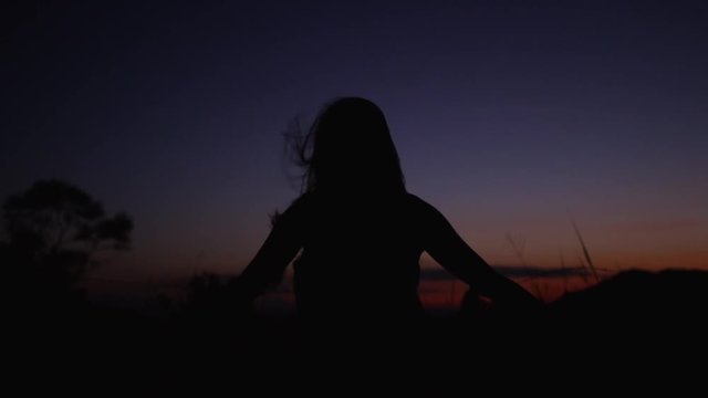 The silhouette of a woman against a vibrant sunset stands with her arms outstretched while a breeze blows her long hair.