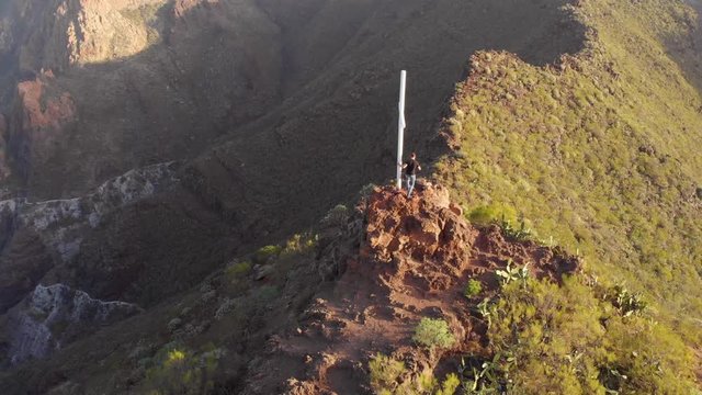 Aerial view of young man climbing up rock and standing near cross admiring epic view. Cruz de Los Misioneros, Tenerife.